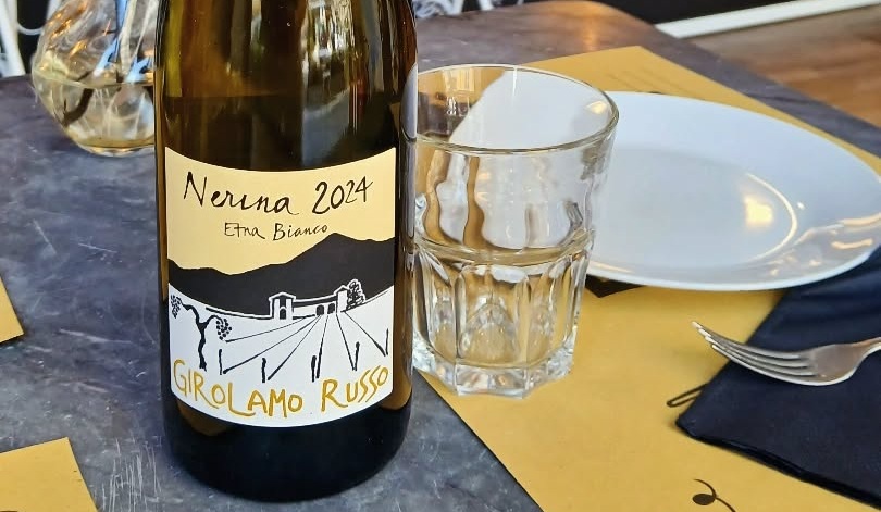 Bottiglia di Etna Bianco Nerina 2024 di Girolamo Russo su tavolo in pietra con bicchiere, piatto bianco e tovaglietta gialla